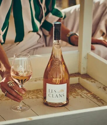Les Clans rosé bottle on a table with a hand holding a glass in a warm setting