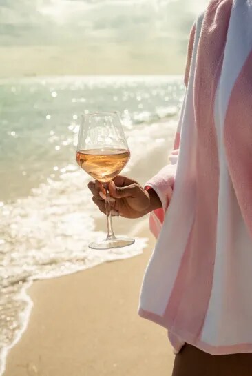 Person holding a glass of rosé on a sunny beach with the sea in the background