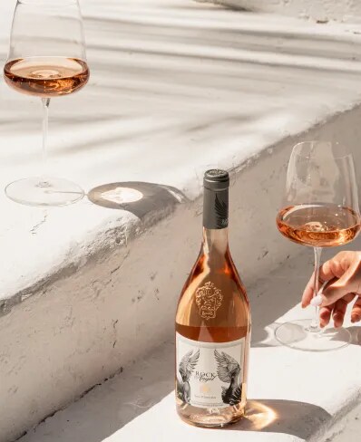 Rock Angel bottle with two glasses on sunlit stone steps, hand holding a glass