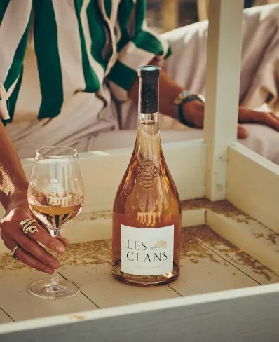 Les Clans rosé bottle on a table with a hand holding a glass in a warm setting